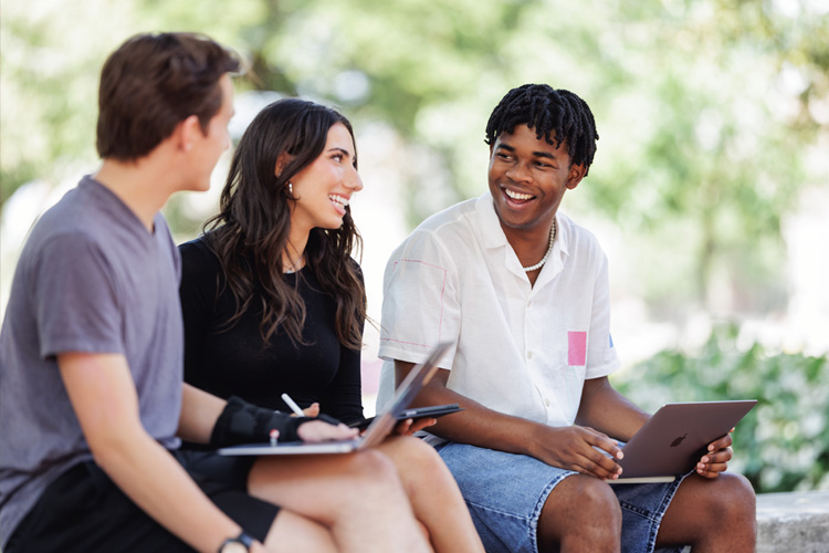 three students talking outdoors on bench with laptops